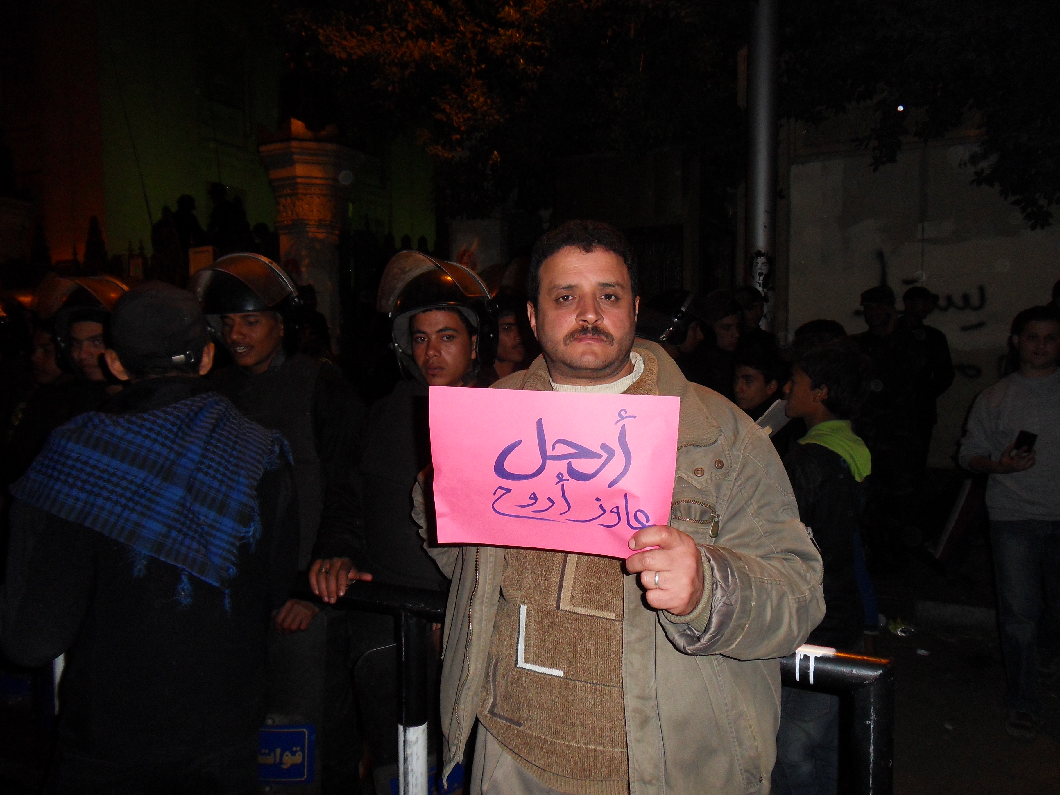 A protesters holding a sign that reads "Leave" while riot police stand behind him guarding the entrance to the Palace.