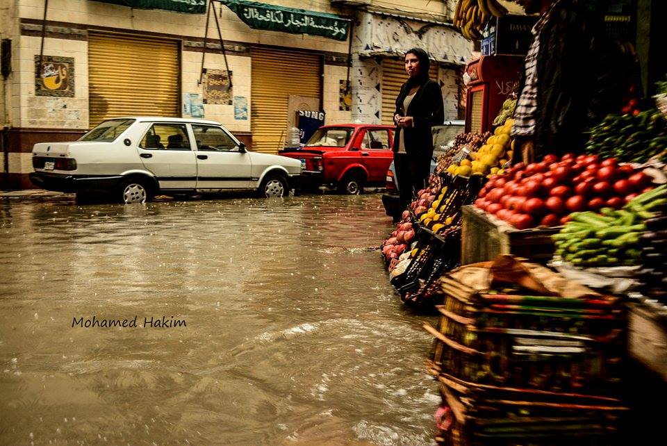 A woman stands on the sidewalk in front of a fruit and vegetable shop, with the rainwater flooding the streets reaching the shop's entrance. Photo: Mohamed Hakim